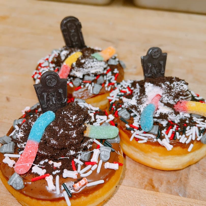 Three Halloween graveyard donuts with cookie-crumb dirt, gummy worms, and tombstone toppers.