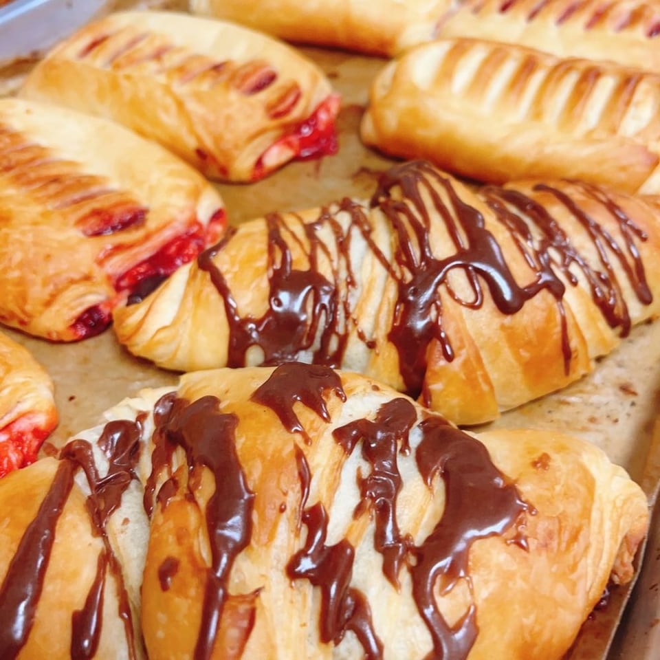 Assortment of pastries on a baking sheet, some with chocolate drizzle, others with red fruit filling.